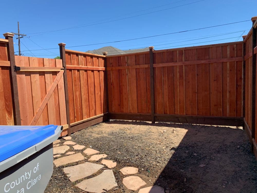 Wooden fenced backyard area with dirt ground and blue trash can in sunny weather.