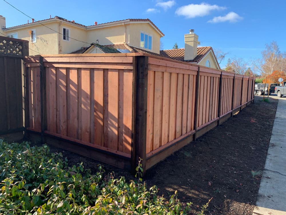 Wooden fence surrounding a landscaped yard with blue sky and houses in the background.