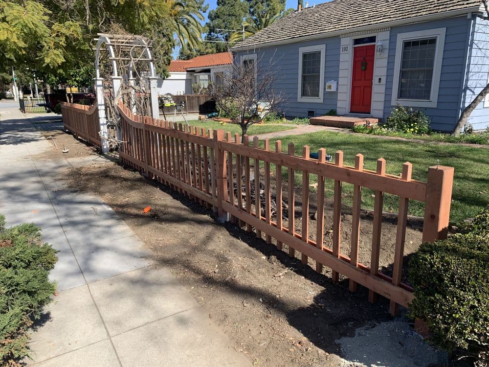 Wooden fence in front of a blue house with red door and landscaped yard.
