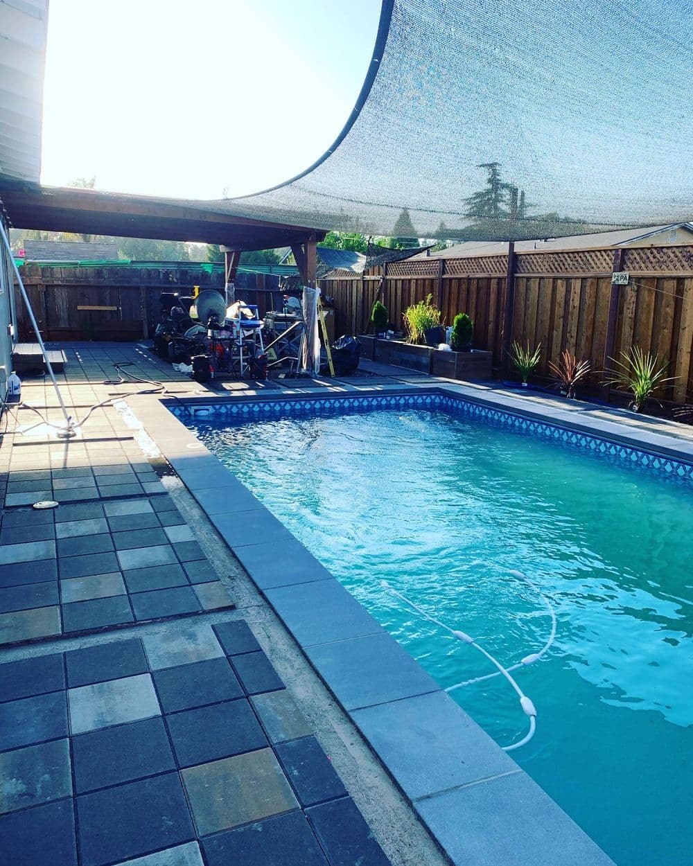 Backyard swimming pool with shaded area and patio, surrounded by landscaping and equipment.
