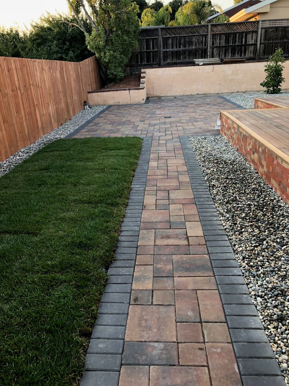 Paved walkway leads through landscaped yard with grass, stones, and wooden fence.