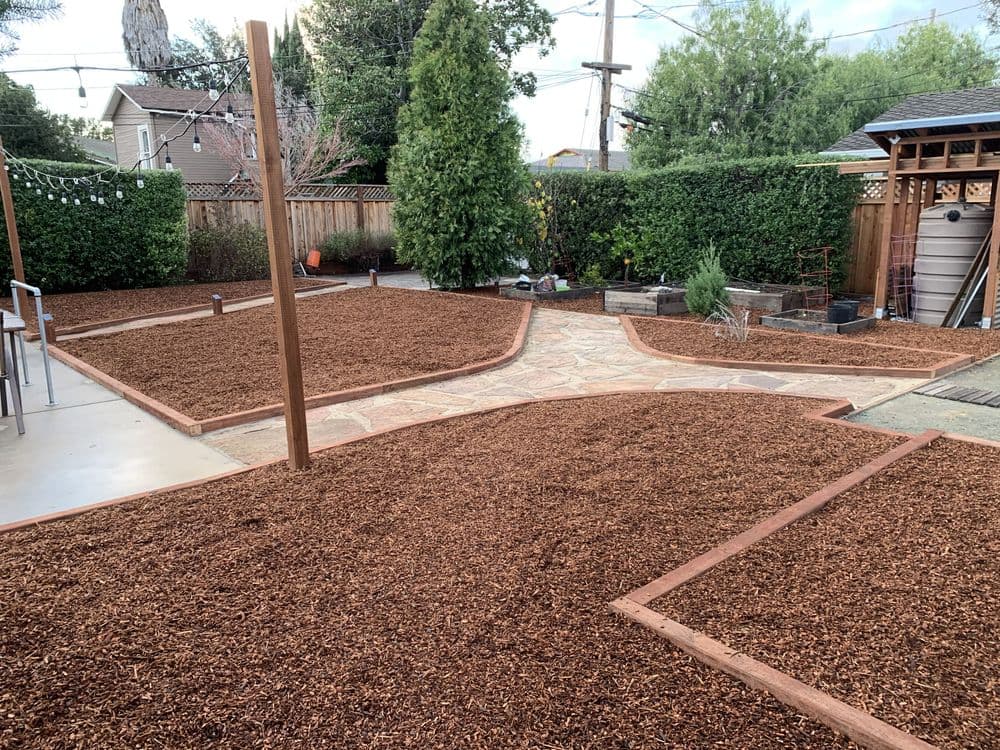 Landscaped garden with stone pathway, wood borders, and mulch beds. Green shrubs in background.