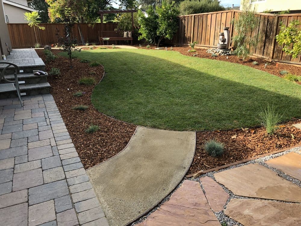 Lush green backyard with stone pathway, landscaped beds, and seating area in a serene setting.
